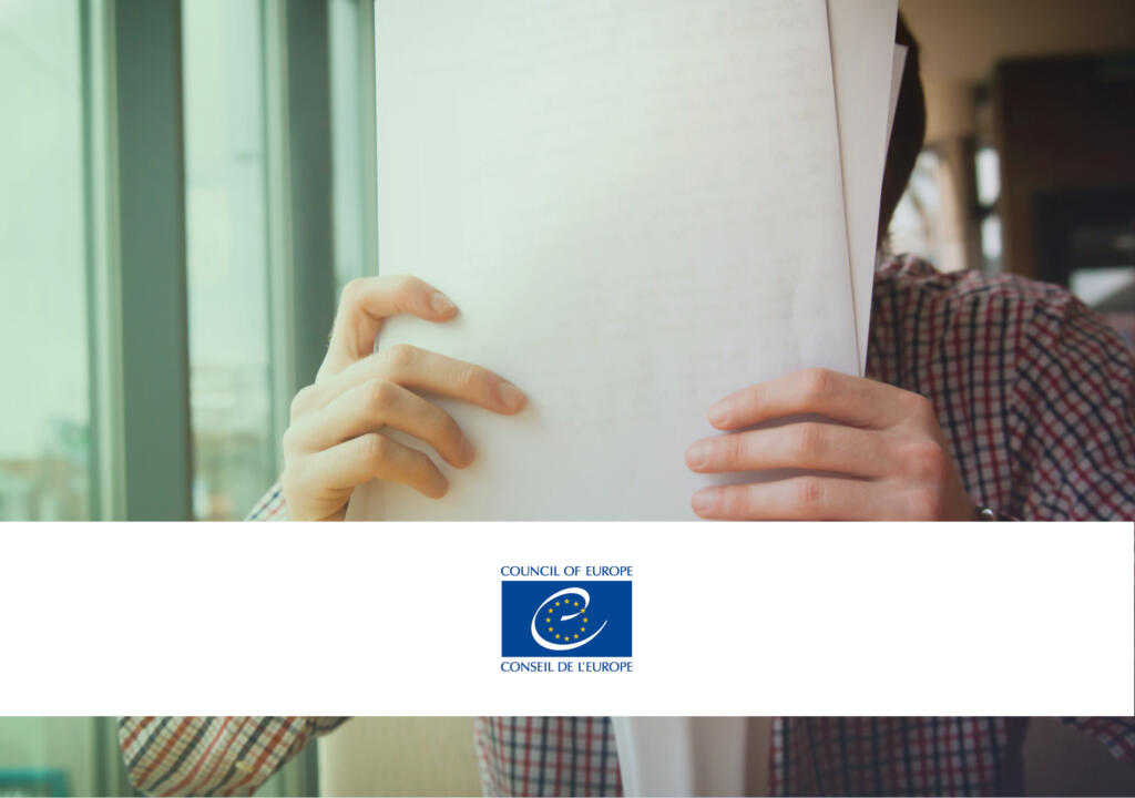 A person holding a pile of paper / a document. There is a white banner with the logo of the Council of Europe, which shows that this policy monitor items is about the Council of Europe.