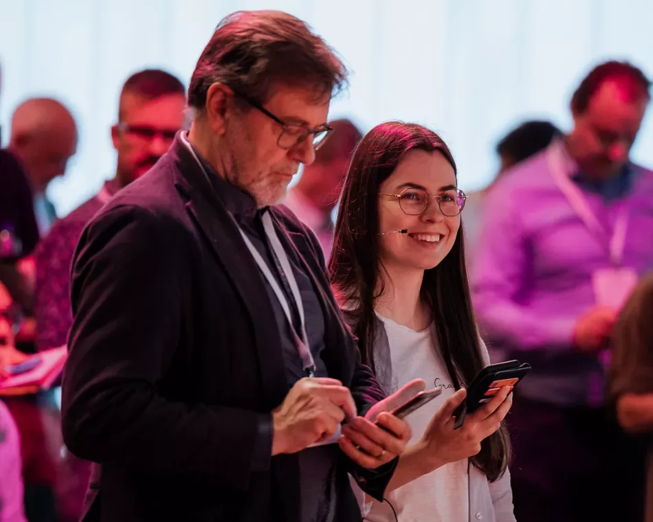 A man in a blazer is looking at his phone. Next to him stands a smiling woman holding a phone. The setting appears to be professional