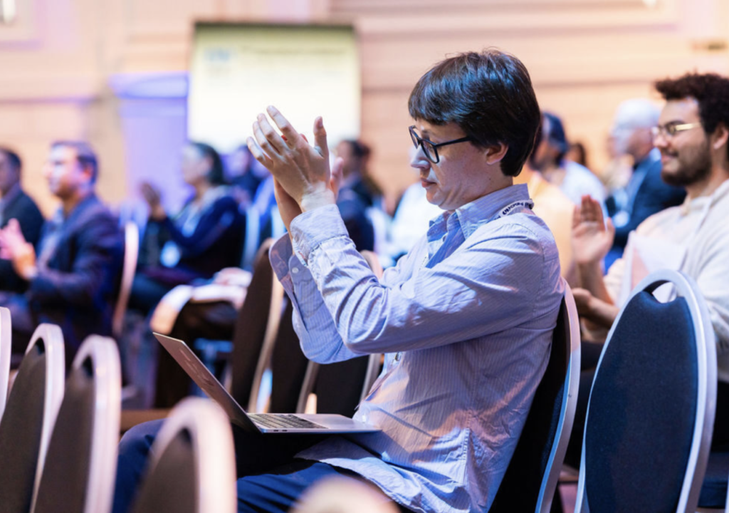 Man op conferentie klapt met zijn handen. Andere personen in de achtergrond applaudisseren ook. 
Man at conference claps his hands. Other people in the background also applaud.