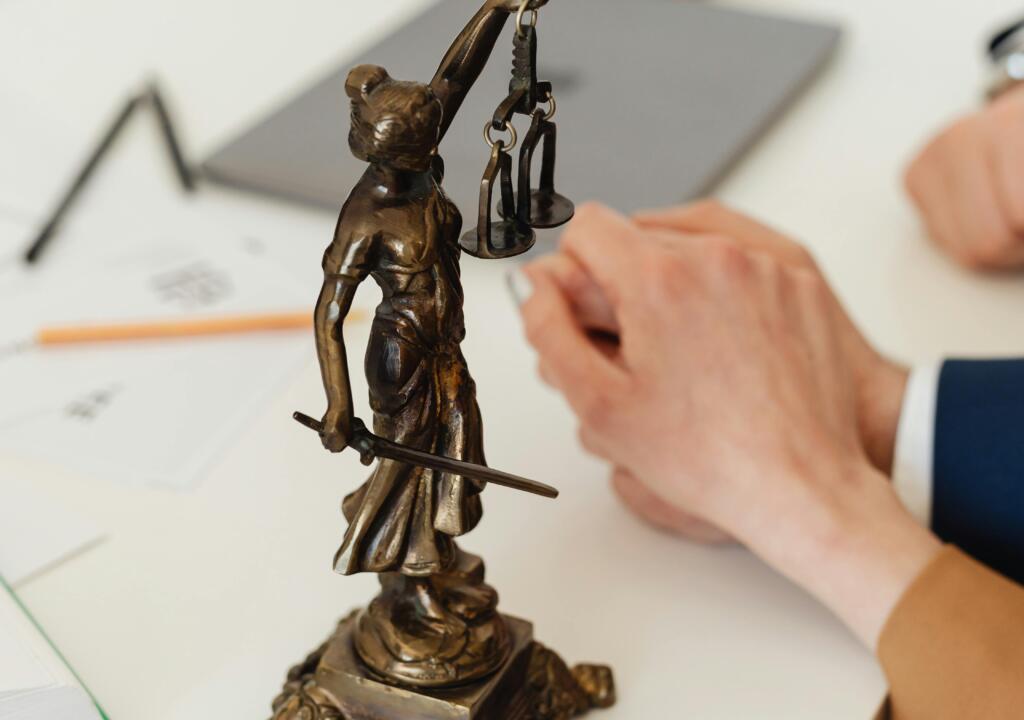 Bronzen beeldje van vrouwe justitia op een bureau. De setting is professioneel. 
Bronze statue of lady justice on a desk. The setting is professional.
