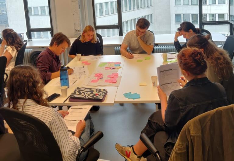 People seated around a table, engaging in a workshop. 
Mensen rond een tafel in een workshop setting.
