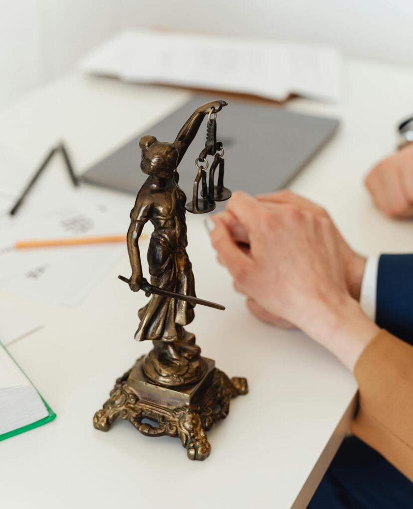 Bronzen beeldje van vrouwe justitia op een bureau. De setting is professioneel. 
Bronze statue of lady justice on a desk. The setting is professional.