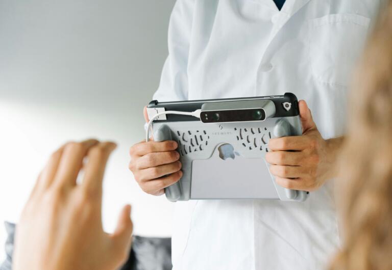 A person in a white lab coat is holding an iPad. The picture shows a medical setting in which the person with the lab coat is making a scan of a patient.