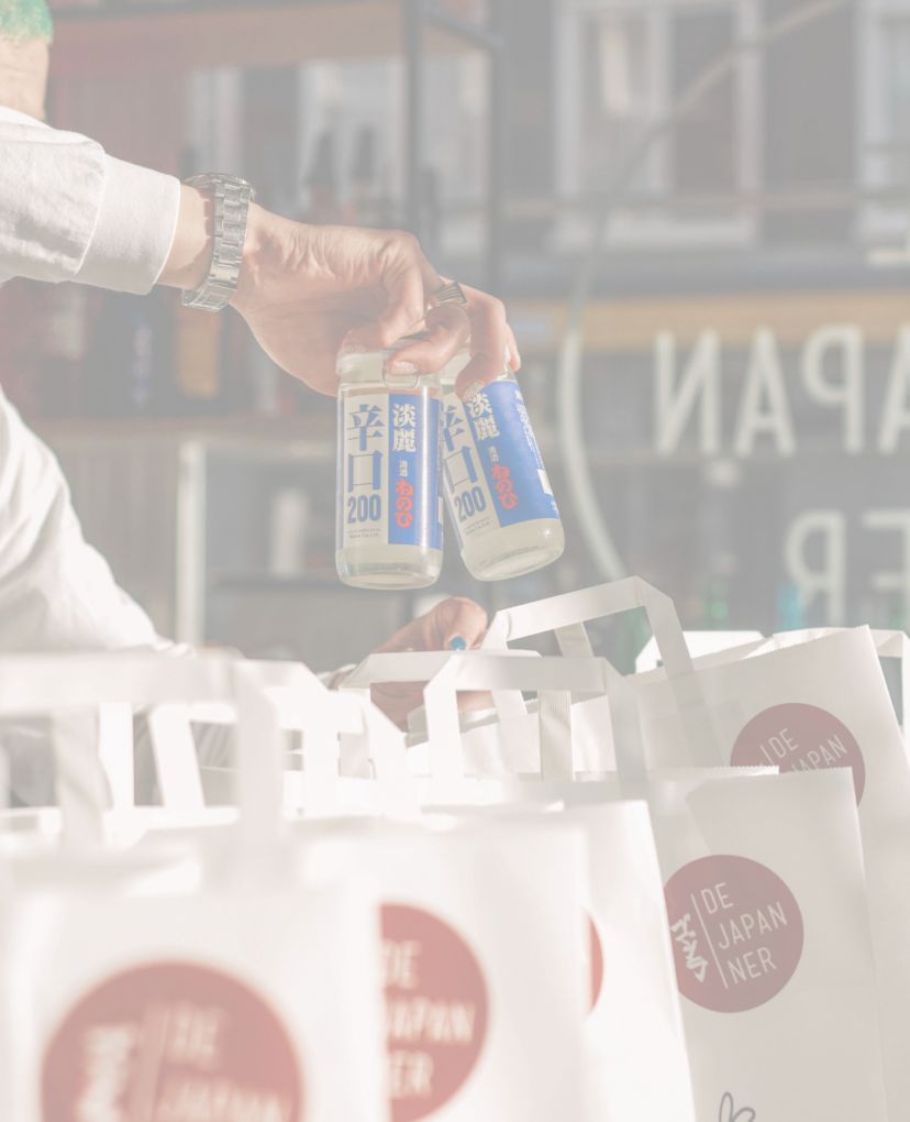 A person holds two canned beverages above several white paper bags with red circular logos. 
Een persoon houdt twee drankblikjes vast boven verschillende papieren tassen met daarop een logo met een rode cirkel.
