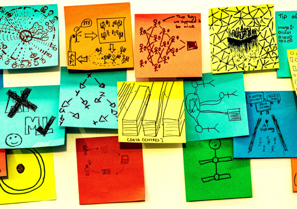 Seventeen multicoloured post-it notes are roughly positioned in a strip shape on a white board. Each one of them has a hand drawn sketch in pen on them, answering the prompt on one of the post-it notes "AI is...." The sketches are all very different, some are patterns representing data, some are cartoons, some show drawings of things like data centres, or stick figure drawings of the people involved.