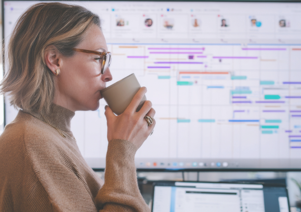 A middle-aged Caucasian woman juggles deadlines while sipping coffee at her standing desk, managing emails, video meetings, and detailed Gantt charts for a high-stakes corporate rollout
