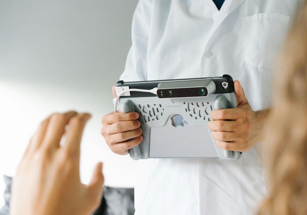 A person in a white lab coat is holding an iPad. The picture shows a medical setting in which the person with the lab coat is making a scan of a patient.