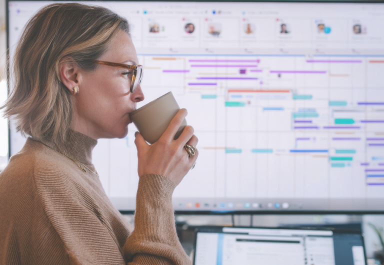 A middle-aged Caucasian woman juggles deadlines while sipping coffee at her standing desk, managing emails, video meetings, and detailed Gantt charts for a high-stakes corporate rollout