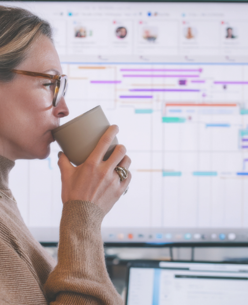 A middle-aged Caucasian woman juggles deadlines while sipping coffee at her standing desk, managing emails, video meetings, and detailed Gantt charts for a high-stakes corporate rollout