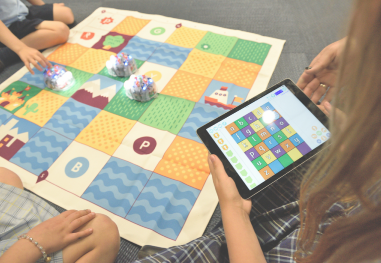 A person uses a tablet while two children enjoy a board game on the floor, smiling and engaged in play.